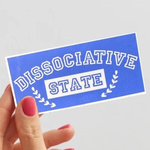 May include: A blue sticker with white text that reads "Dissociative State" with a white wreath design on either side of the text.