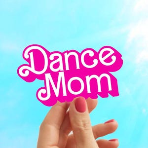 May include: A hand holds up a pink sign with the words "Dance Mom" in white letters.