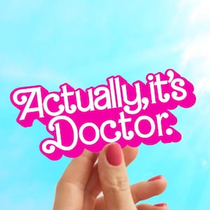 May include: A pink sign with white text that reads "Actually, it's Doctor." The sign is being held up against a blue sky.