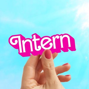May include: A hand holds up a pink sign with the word "Intern" written in white letters.