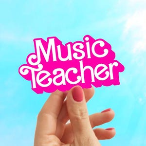 May include: A hand holds up a pink sign with the words "Music Teacher" in white letters against a blue sky.