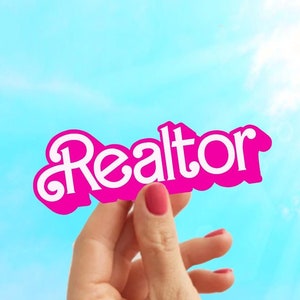 May include: A hand holding up a pink sign with the word "Realtor" in white letters against a blue sky.