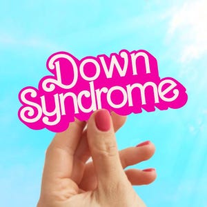 May include: A hand holds up a pink sign with the words "Down Syndrome" in white letters.