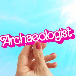 May include: A pink and white sign that reads "Archaeologist" is held up against a bright blue sky with the sun shining. The sign is held by a hand with red nail polish.