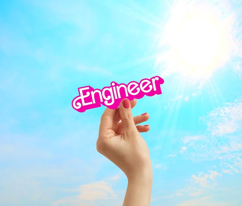 May include: A hand holds up a pink paper sign with the word "Engineer" written in white letters against a bright blue sky.