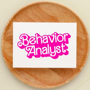 May include: A white card with the words "Behavior Analyst" in bright pink, stylized lettering. The card is placed on a round, wooden tray with a light brown color. The background is a neutral beige.