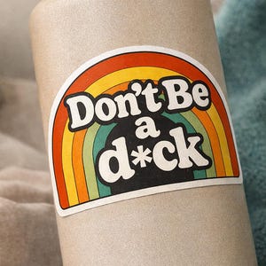May include: A beige water bottle with a black lid and handle. A sticker on the bottle features a rainbow design with the words "Don't Be a d*ck" in bold, white letters.