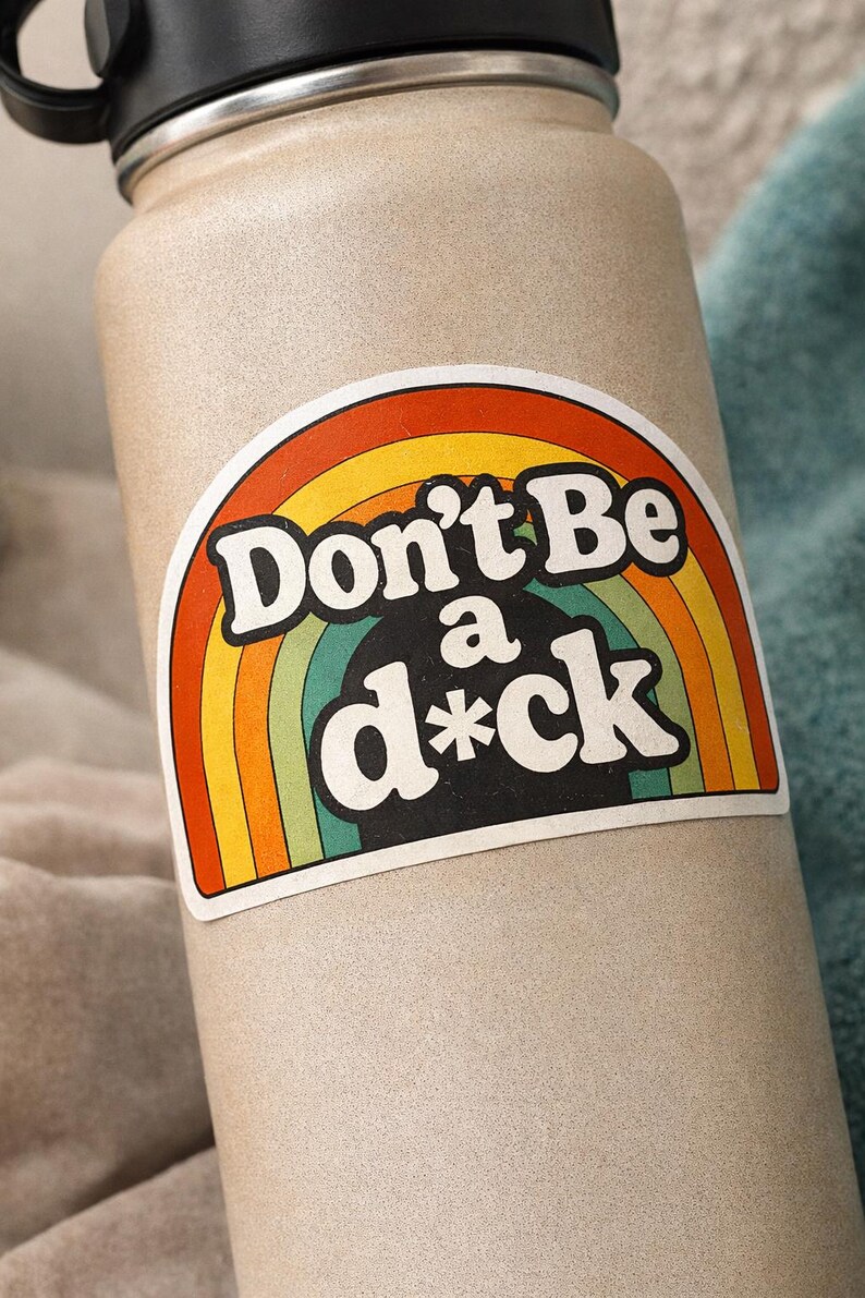 May include: A beige water bottle with a black lid and handle. A sticker on the bottle features a rainbow design with the words "Don't Be a d*ck" in bold, white letters.