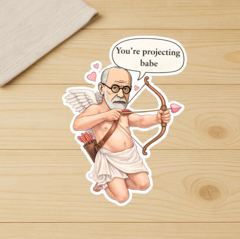 May include: A sticker featuring a cartoon illustration of Sigmund Freud as Cupid, holding a bow and arrow. The speech bubble says, "You're projecting babe." The sticker has a white border and is on a wooden surface.