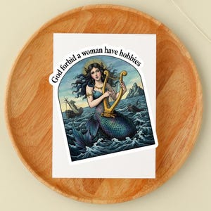 May include: A sticker featuring a mermaid playing a golden harp in a sea scene with ships. The mermaid has long dark hair and a halo. The sticker has the text "God forbid a woman have hobbies" in a circular design.