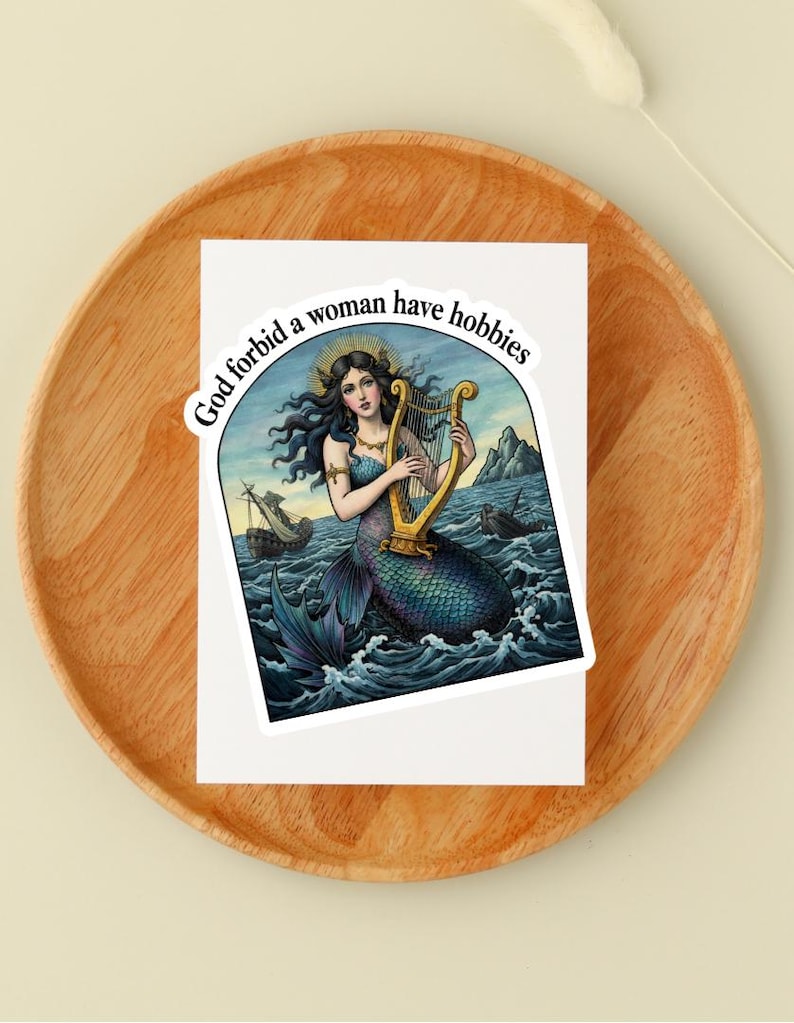 May include: A sticker featuring a mermaid playing a golden harp in a sea scene with ships. The mermaid has long dark hair and a halo. The sticker has the text "God forbid a woman have hobbies" in a circular design.
