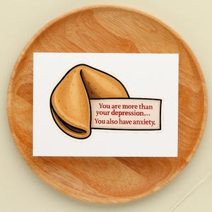 May include: A white card with a fortune cookie illustration and the text: "You are more than your depression... You also have anxiety." The card is on a wooden plate.