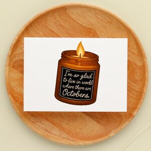 May include: A greeting card featuring a candle in a brown jar with a lit orange flame. The jar has a black label with the text "I'm so glad to live in world where there are Octobers."