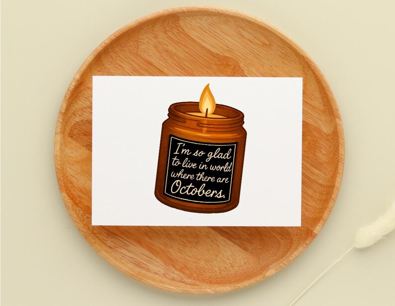 May include: A greeting card featuring a candle in a brown jar with a lit orange flame. The jar has a black label with the text "I'm so glad to live in world where there are Octobers."