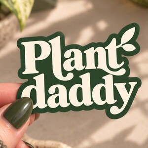 May include: A die-cut sticker featuring the text Plant daddy in a bold, cream-coloured serif font against a dark forest green background. The design includes a small leaf graphic attached to the letter y, with a thick green border.