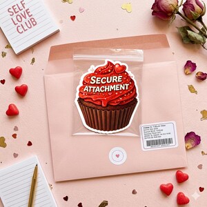 May include: A cupcake sticker with red icing and the words "SECURE ATTACHMENT" in a clear plastic bag, inside a pink envelope. The background includes a notepad that says "SELF LOVE CLUB", heart-shaped sweets, and dried roses.