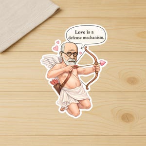 May include: A sticker featuring a cartoon of Sigmund Freud as Cupid, with wings and a bow and arrow. The speech bubble says, "Love is a defense mechanism." The sticker is on a wooden surface.