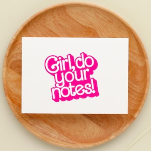 May include: A white card with the phrase "Girl, do your notes!" in vibrant pink lettering. The card rests on a wooden plate. The font is a playful, bubble-style design.
