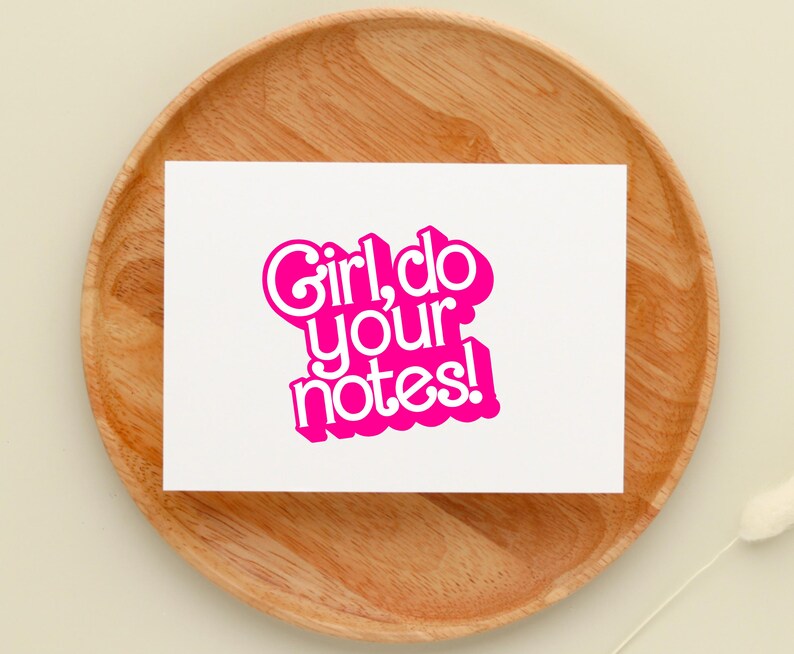 May include: A white card with the phrase "Girl, do your notes!" in vibrant pink lettering. The card rests on a wooden plate. The font is a playful, bubble-style design.