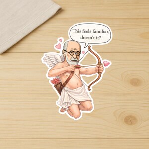 May include: A sticker featuring a cartoon of Sigmund Freud as Cupid, with wings, a bow and arrow, and a quiver of arrows. The speech bubble says, "This feels familiar, doesn't it?" The sticker is on a wooden surface.