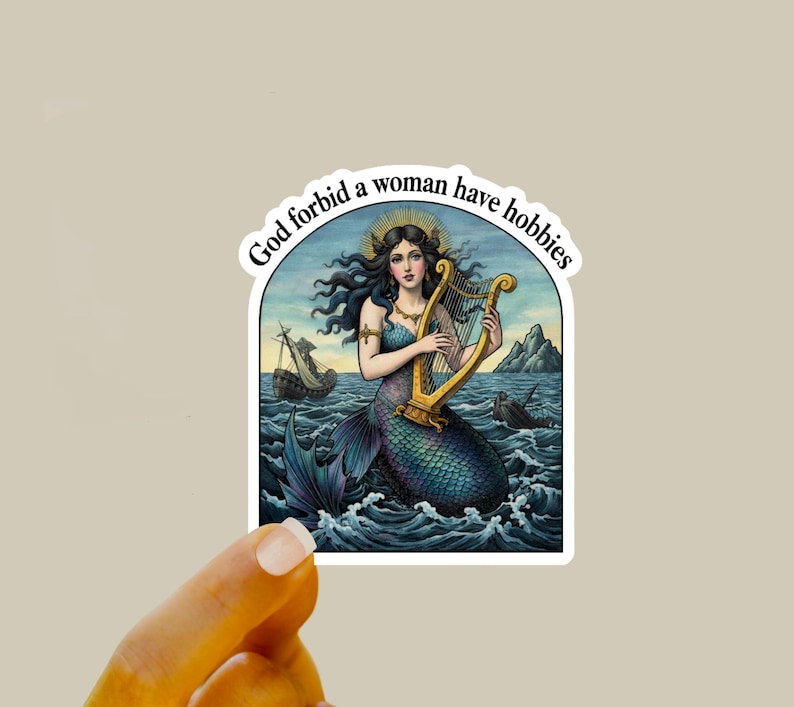 May include: A sticker featuring a mermaid playing a golden harp in the ocean. The mermaid has long dark hair and a blue tail. The sticker has the text "God forbid a woman have hobbies" in a white arch.
