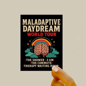 Op de afbeelding: Zwarte sticker met de tekst "MALADAPTIVE DAYDREAM WORLD TOUR" in een retro lettertype. Het ontwerp bevat een hersen met een koptelefoon met een regenboogboog, wolken en sterren. Hieronder staat "THE SHOWER, 3 AM, THE COMMUTE, THERAPY WAITING ROOM."