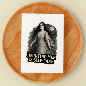 May include: A sticker featuring a vintage illustration of a woman with long, flowing hair and a flowing gown. The sticker has the text "Haunting Men Is Self-Care" in a bold, black font. The sticker is placed on a wooden plate.