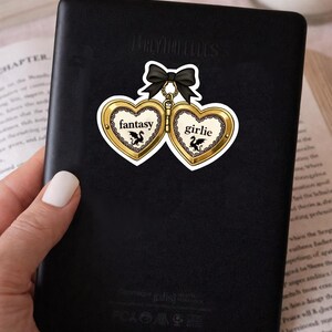 May include: A black sticker featuring two heart-shaped lockets with the words "fantasy" and "girlie" in gold lettering. Each locket has a black bow and a small illustration of a dragon. The sticker is on a black surface.