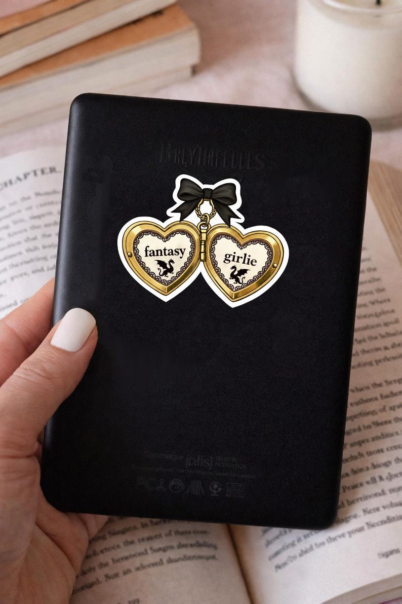 May include: A black sticker featuring two heart-shaped lockets with the words "fantasy" and "girlie" in gold lettering. Each locket has a black bow and a small illustration of a dragon. The sticker is on a black surface.