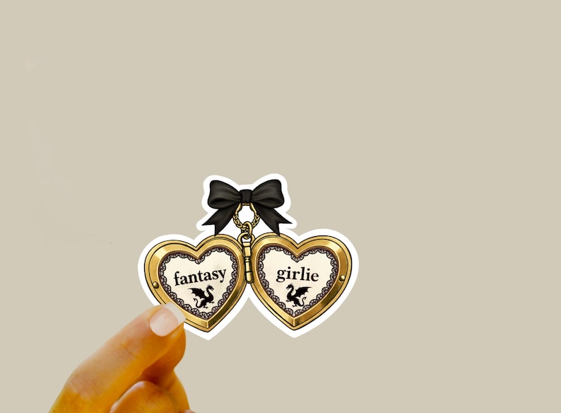 May include: A gold heart-shaped pendant with the words "fantasy" and "girlie" on each side. Each heart has a black dragon illustration. The pendant is connected by a black bow.