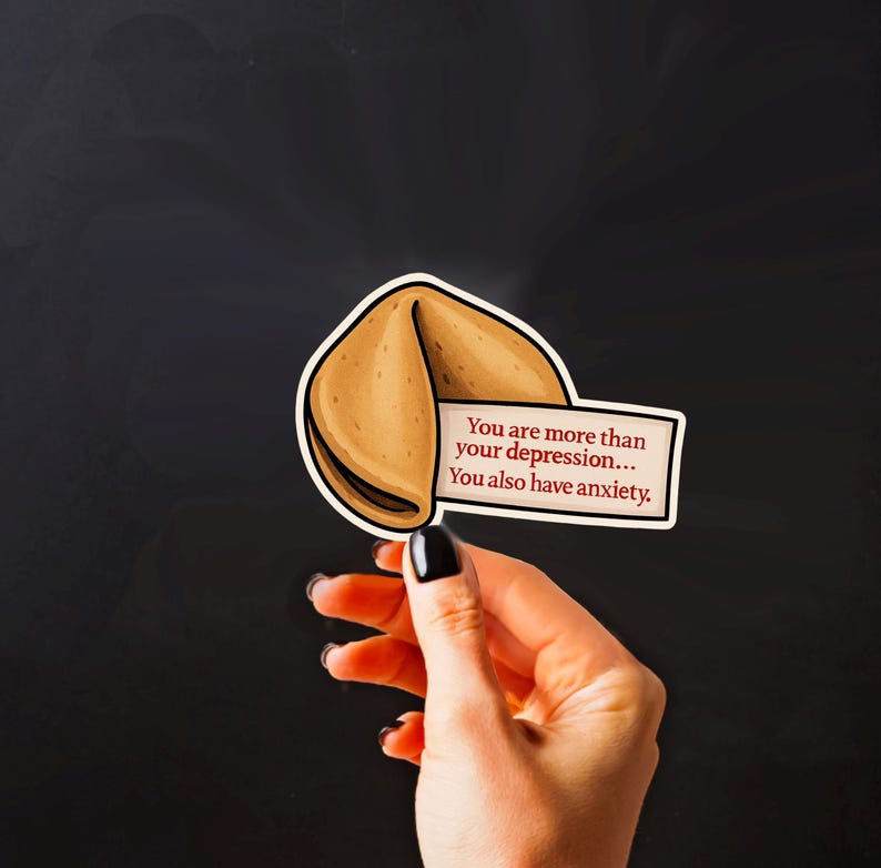 May include: A sticker in the shape of a fortune cookie, with the text "You are more than your depression... You also have anxiety." The cookie is light brown with a white border, and the text is on a white rectangle.