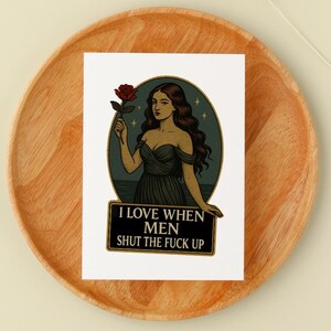 May include: A sticker featuring a woman holding a red rose, set against a teal background with stars. The text "I LOVE WHEN MEN SHUT THE FUCK UP" is displayed on a black banner below the illustration. The sticker is placed on a wooden plate.