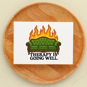 May include: A white sticker featuring a green couch engulfed in flames, with the text "THERAPY IS GOING WELL." The couch has a tufted back and wooden frame. The flames are orange and yellow.