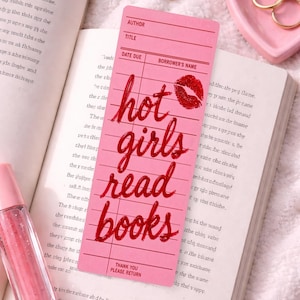 May include: Pink bookmark with the text "hot girls read books" in red script, a red lip print, and a book. A pink heart-shaped dish, gold rings, and a lip gloss are also visible.