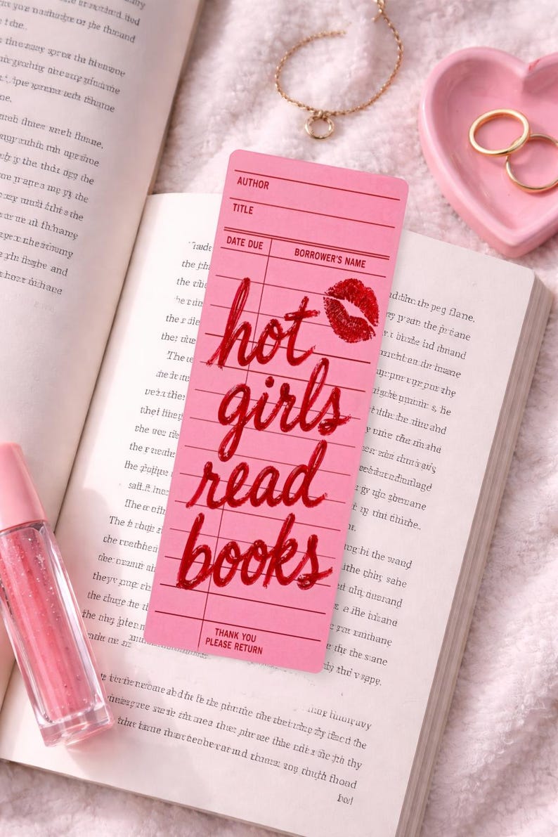 May include: Pink bookmark with the text "hot girls read books" in red script, a red lip print, and a book. A pink heart-shaped dish, gold rings, and a lip gloss are also visible.
