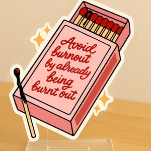 May include: A pink matchbox-shaped acrylic stand with the text "Avoid burnout by already being burnt out." The matchbox contains red-tipped matches. A burnt match is next to the box. The stand is on a clear base.
