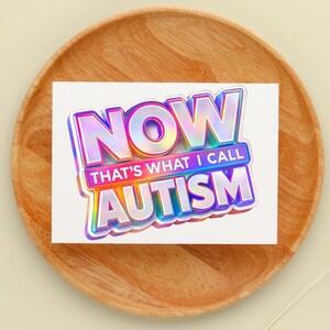 May include: A white card with the text "NOW THAT'S WHAT I CALL AUTISM" in a holographic, 3D-style font. The card is on a wooden plate, with a light beige background.