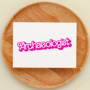 May include: A white card with the word "Archaeologist" in a bright pink, bubble-style font. The card is set on a round, wooden tray with a light-colored background. A decorative element is visible in the lower right corner.