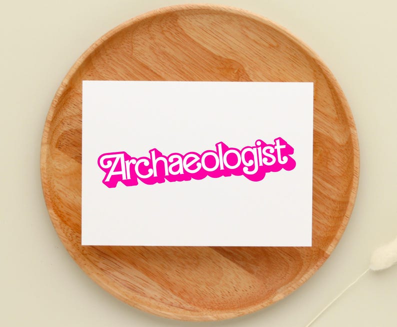 May include: A white card with the word "Archaeologist" in a bright pink, bubble-style font. The card is set on a round, wooden tray with a light-colored background. A decorative element is visible in the lower right corner.