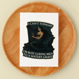 May include: A white sticker featuring a mermaid playing a harp. The mermaid has long dark hair and a green tail. Text on the sticker reads "I CAN'T TONIGHT" and "I'M BUSY LURING MEN TO A WATERY GRAVE."