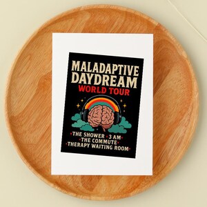 Op de afbeelding: Een witte kaart met de tekst "MALADAPTIVE DAYDREAM WORLD TOUR" in zwart en rood. De kaart toont een hersengrafiek met een koptelefoon, een regenboog en de zinnen "THE SHOWER 3 AM", "THE COMMUTE" en "THERAPY WAITING ROOM". De kaart ligt op een houten bord.