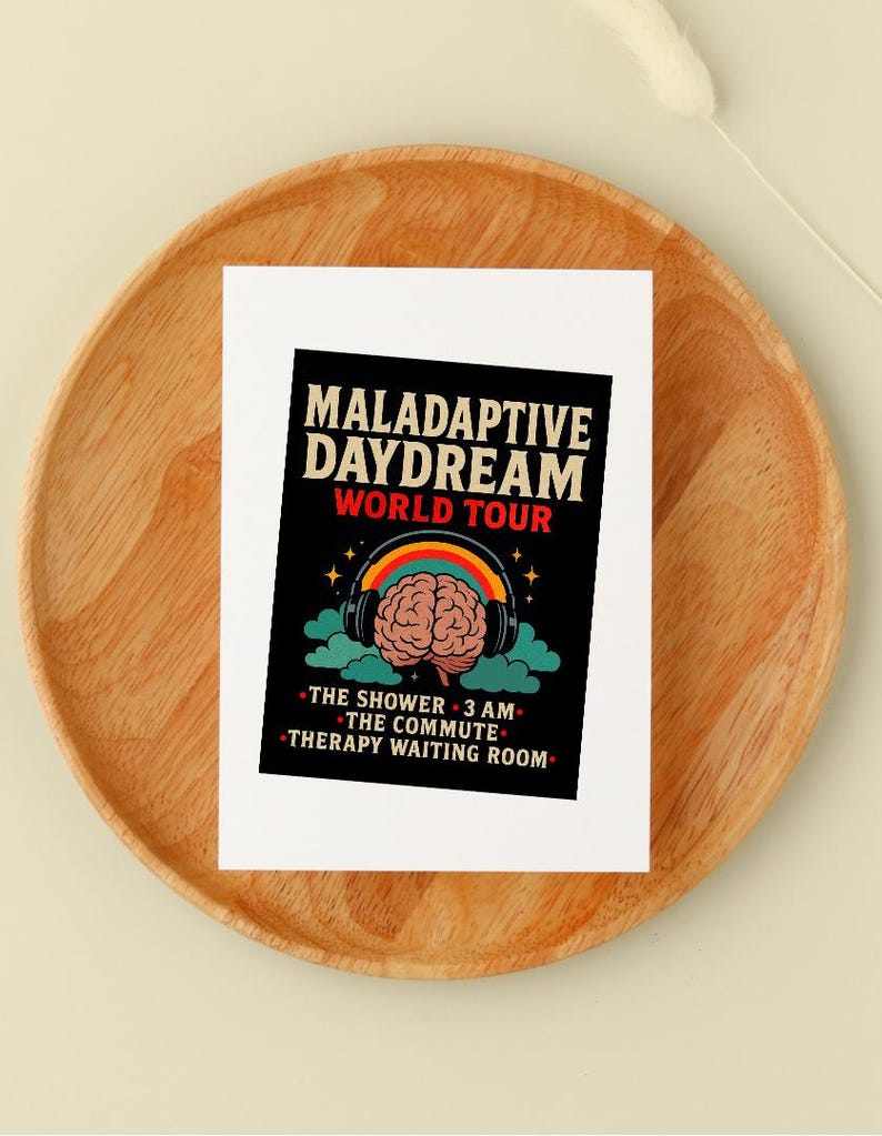 Op de afbeelding: Een witte kaart met de tekst "MALADAPTIVE DAYDREAM WORLD TOUR" in zwart en rood. De kaart toont een hersengrafiek met een koptelefoon, een regenboog en de zinnen "THE SHOWER 3 AM", "THE COMMUTE" en "THERAPY WAITING ROOM". De kaart ligt op een houten bord.