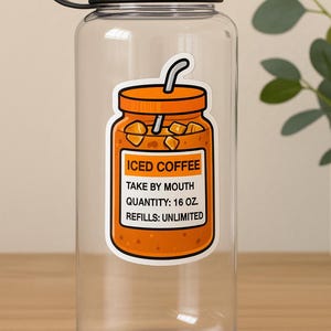 Peut inclure: Une bouteille d'eau transparente avec un couvercle et une poign&eacute;e noirs. Un autocollant orange de caf&eacute; glac&eacute; est appos&eacute; sur la bouteille, avec le texte "ICED COFFEE, TAKE BY MOUTH, QUANTITY: 473 ml, REFILLS: UNLIMITED."