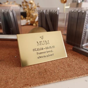 May include: A gold-colored rectangular plaque with the word "MUM" and the dates 07.12.59-13.01.15, along with the words "Forever loved. always missed." Two small hearts are above the word "MUM".