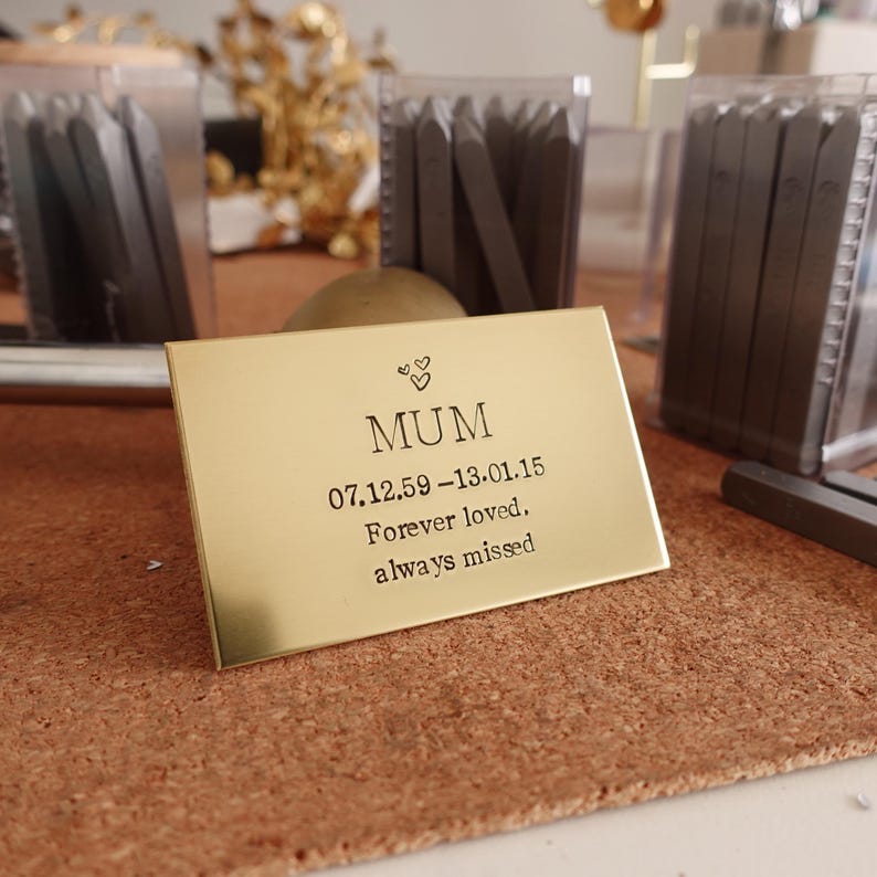 May include: A gold-colored rectangular plaque with the word "MUM" and the dates 07.12.59-13.01.15, along with the words "Forever loved. always missed." Two small hearts are above the word "MUM".