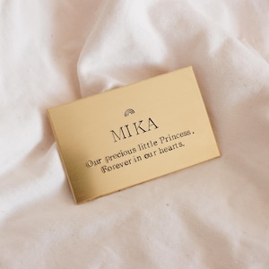 May include: A rectangular gold-colored plaque with the name "MIKA" and the phrase "Our precious little Princess. Forever in our hearts." The plaque has a small rainbow design above the name and is set against a soft, white fabric background.