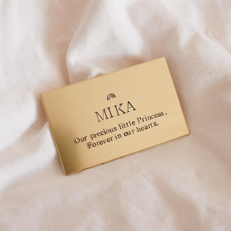 May include: A rectangular gold-colored plaque with the name "MIKA" and the phrase "Our precious little Princess. Forever in our hearts." The plaque has a small rainbow design above the name and is set against a soft, white fabric background.