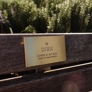 May include: A gold-colored plaque with the name "DIRK" and the inscription "Always on our laps, now in our hearts." The plaque is affixed to a wooden bench, with a blurred green background of foliage.