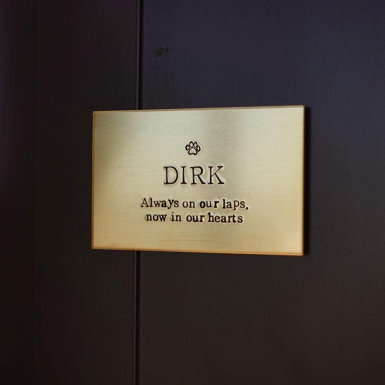 May include: A rectangular brass plaque with the name "DIRK" and a paw print symbol. The text below reads "Always on our laps, now in our hearts". The plaque is mounted on a dark brown surface.