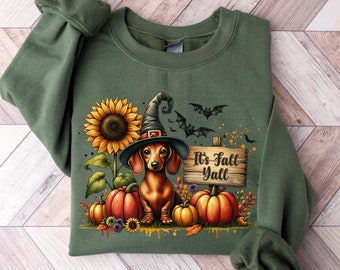 May include: An olive green crewneck sweatshirt featuring a graphic of a dachshund wearing a witch hat, surrounded by sunflowers, pumpkins, and bats. A wooden sign next to the dog displays the text It's Fall Yall in black script lettering.